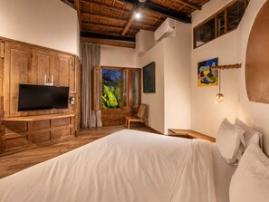 Une chambre avec un meuble TV en bois, un lit, une fenêtre donnant sur un paysage verdoyant et un tableau accroché au mur.