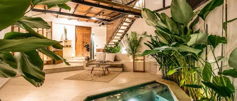 An indoor swimming pool with a glass barrier, surrounded by large green plants, a wooden staircase, and a seating area with a sofa and coffee table.