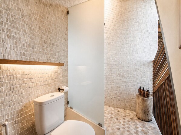 Une salle de bain avec des toilettes blanches, une douche et un mur carrelé sur lequel se trouve une étagère en bois au-dessus des toilettes.