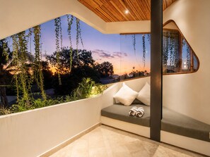 Een modern balkon met een gebogen glazen wand, een zitgedeelte met kussens en uitzicht op een zonsondergang.