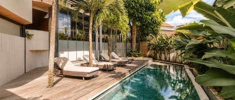 Une piscine entourée de chaises longues, de plantes tropicales et d’un bâtiment moderne.