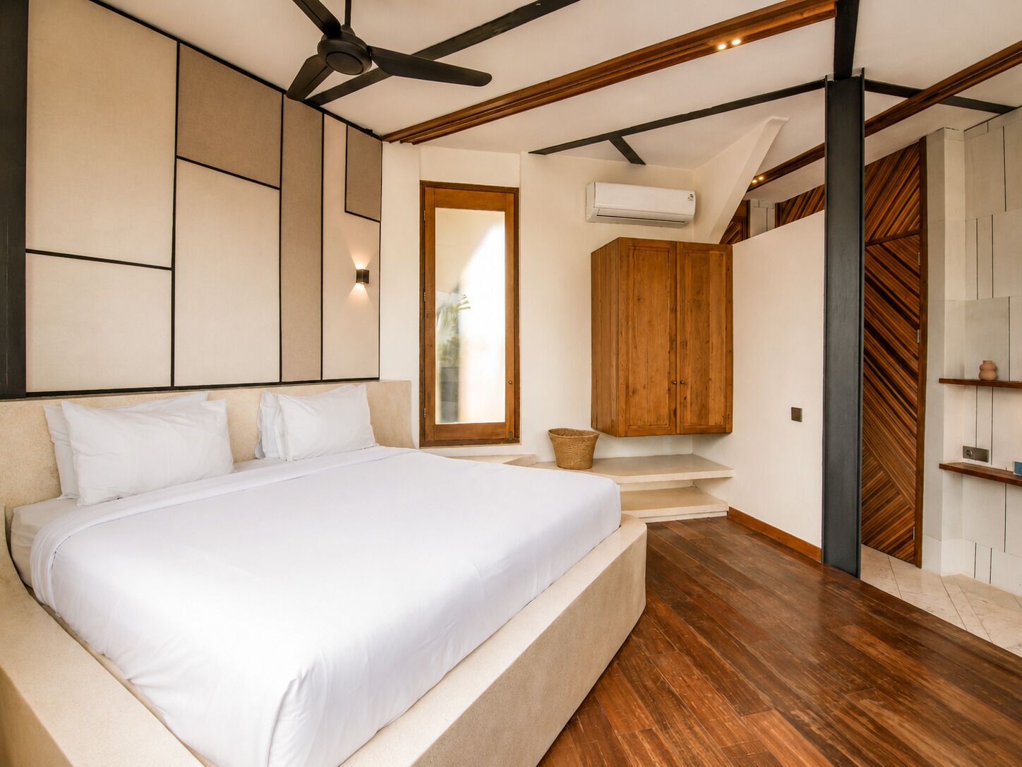 A modern bedroom with a large bed, wooden ceiling beams, an air conditioning unit, and a wooden wardrobe.