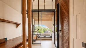 Un interior moderno con un escritorio de madera, una zona de estar con cojines y vistas, y un puertas corredera que da a un espacio exterior.