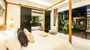 A modern bedroom with a large bed, a chair, and a view of a lush garden through glass doors.
