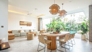 Amplia sala de estar con una mesa redonda de madera, asientos cómodos y un ventanal con vistas a un jardín exuberante y una piscina.