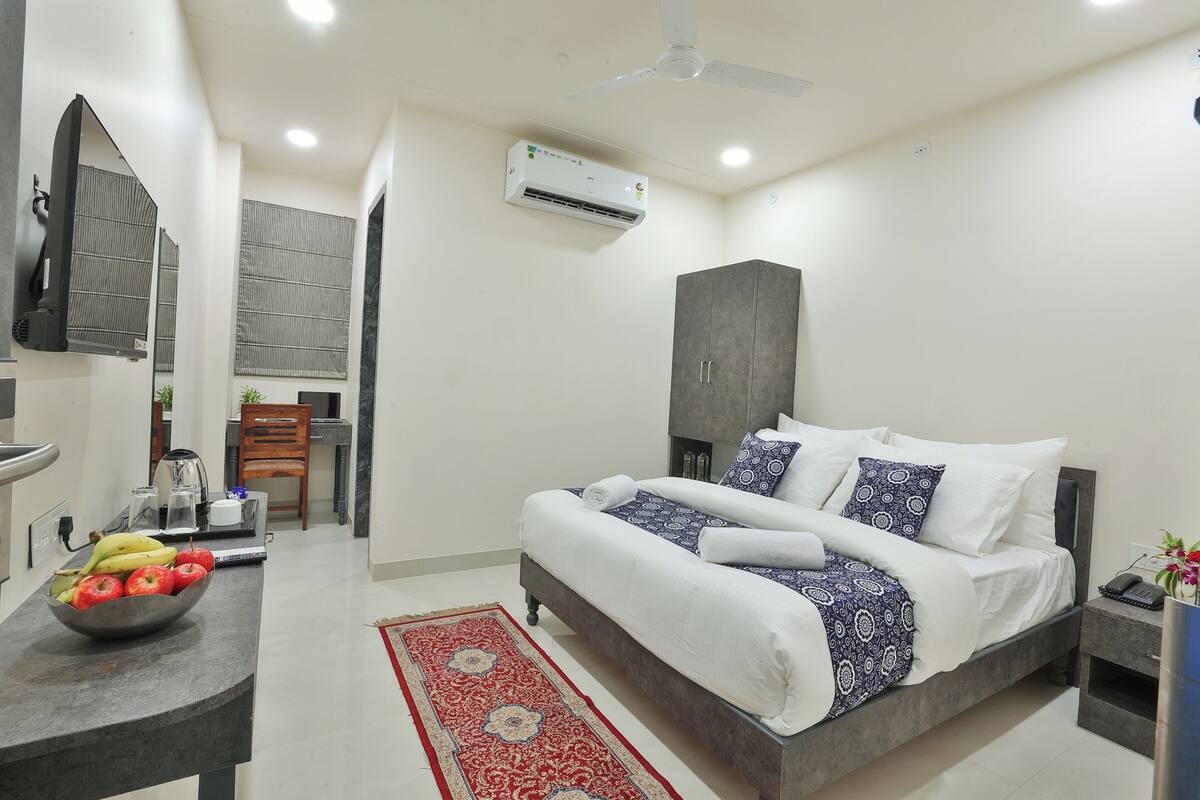 A modern hotel room with a bed, a desk with a fruit bowl, a wardrobe, and an air conditioning unit.