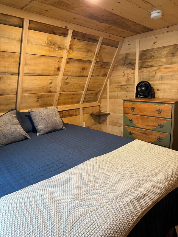 A bedroom with a sloped ceiling, a bed with a dark blue bedspread, two pillows, a green dresser with drawers, and a ceiling fan. - Sister A-Frame in Woods B (Oakland)