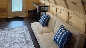 A cozy attic room with wooden walls, a ceiling fan, a window with blinds, a sofa with patterned cushions, and a small table. - Sister A-Frame in Woods B (Oakland)