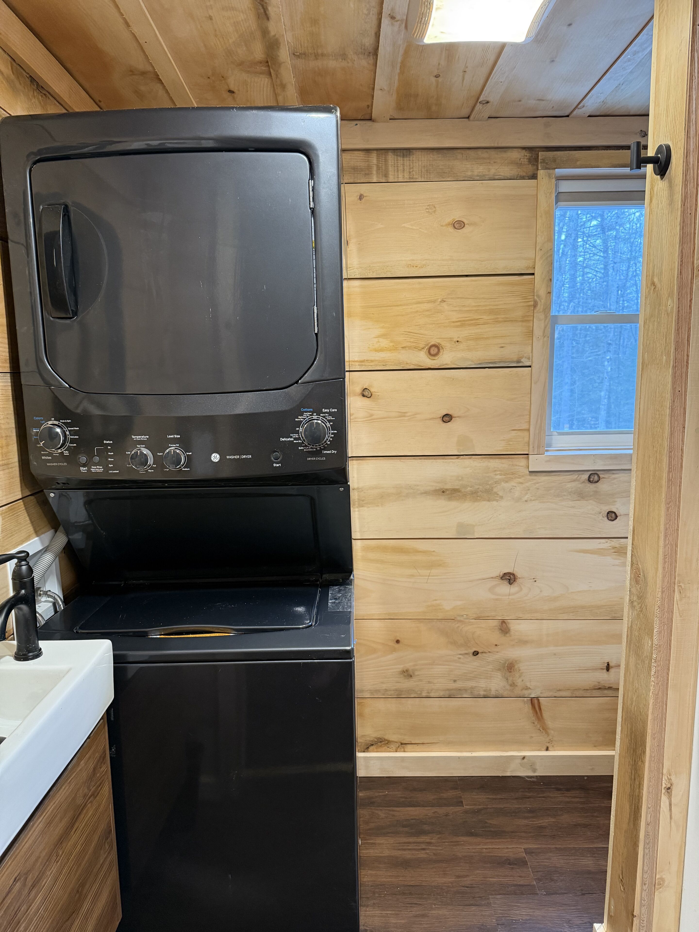 A compact kitchen with a black washing machine, wooden walls, a window, and a sink.