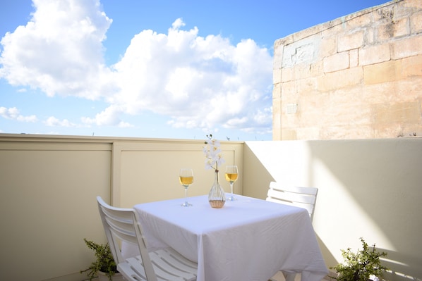 A table set with two glasses of white wine and a vase with white flowers, surrounded by white chairs, against a beige wall under a clear sky.