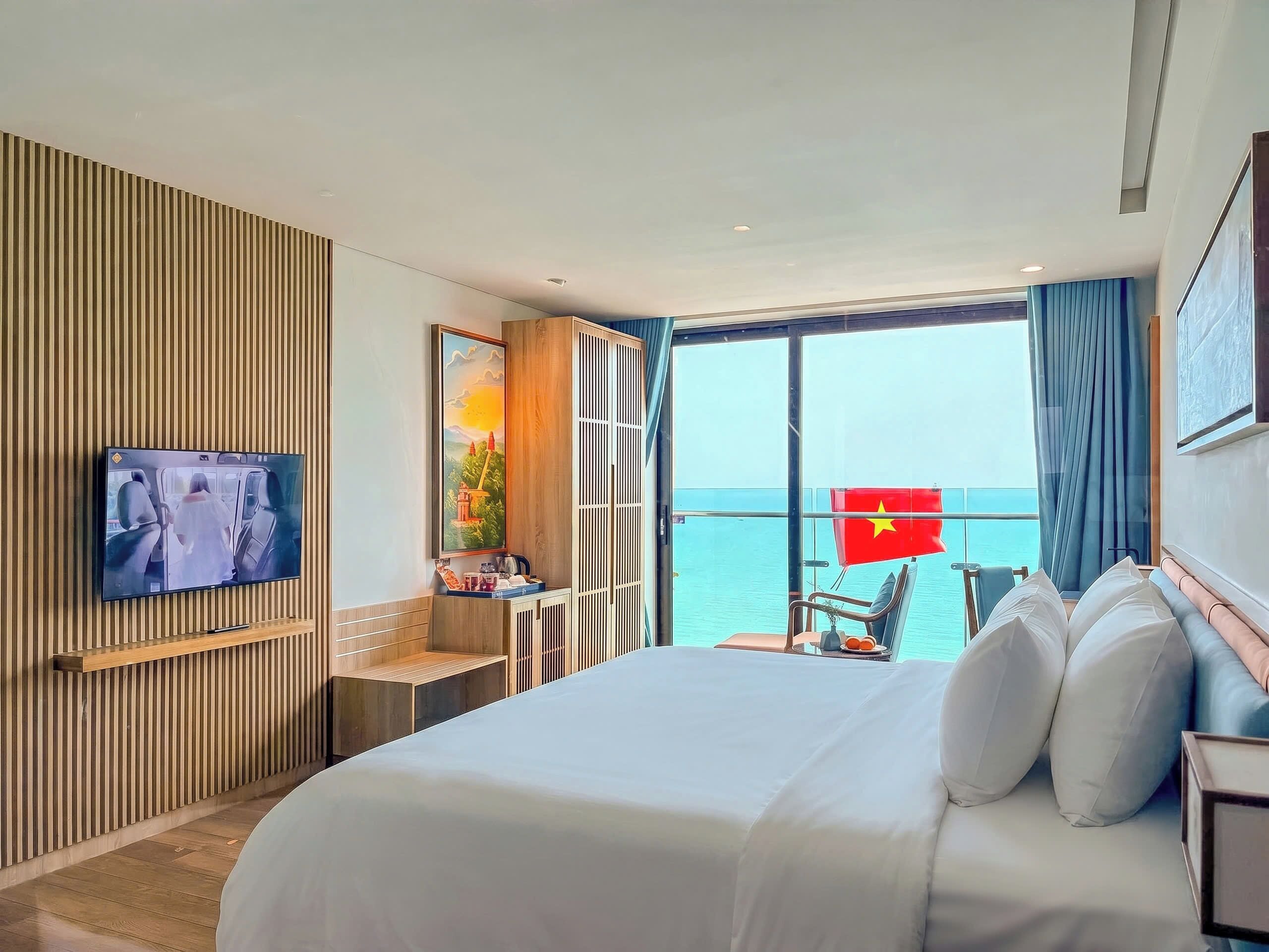 Habitación de hotel con cama, televisor, escritorio con silla, balcón con una bandera y vista al mar.