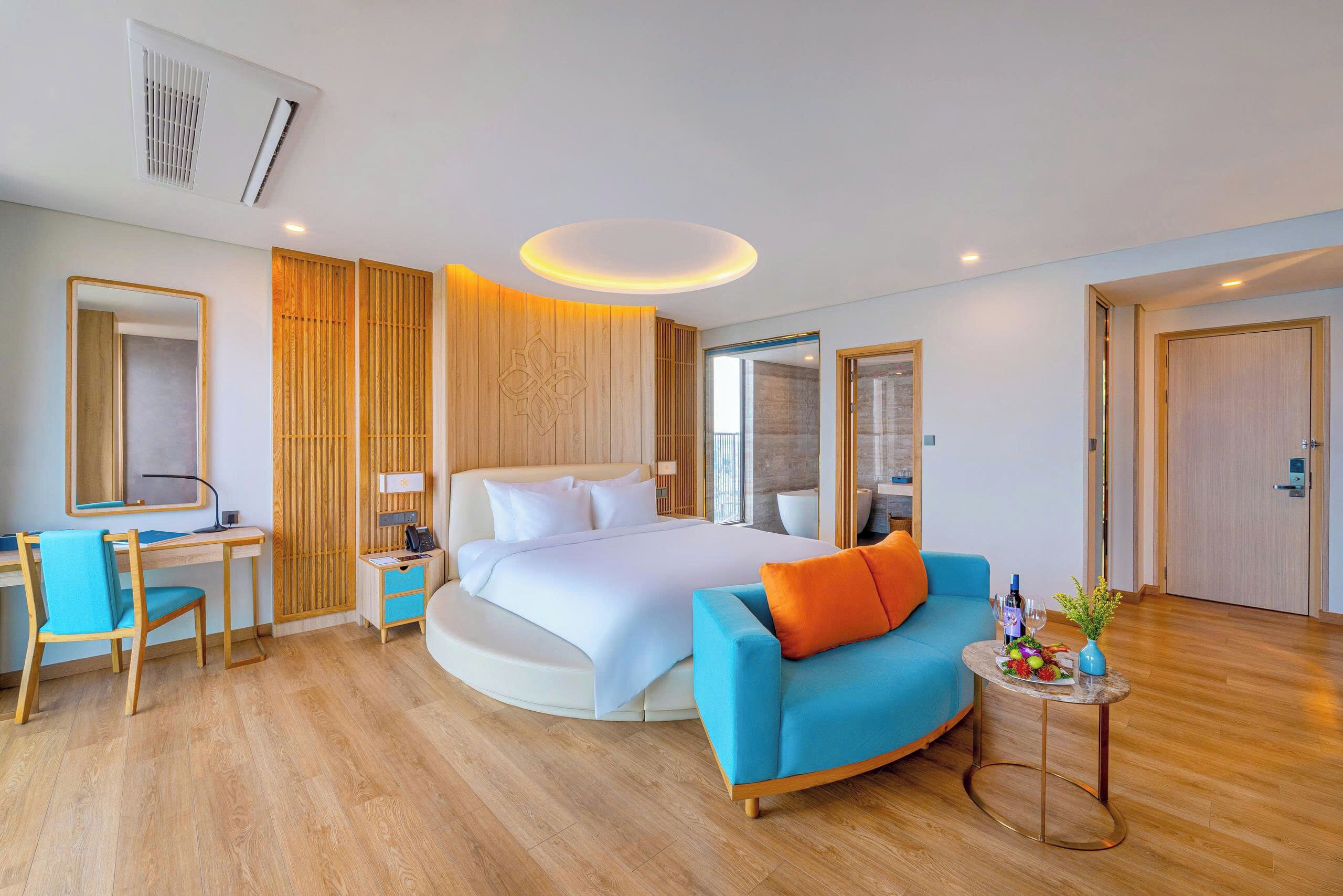 A modern hotel room with a bed, a desk, a chair, a sofa, and a small table with a vase and fruit.
