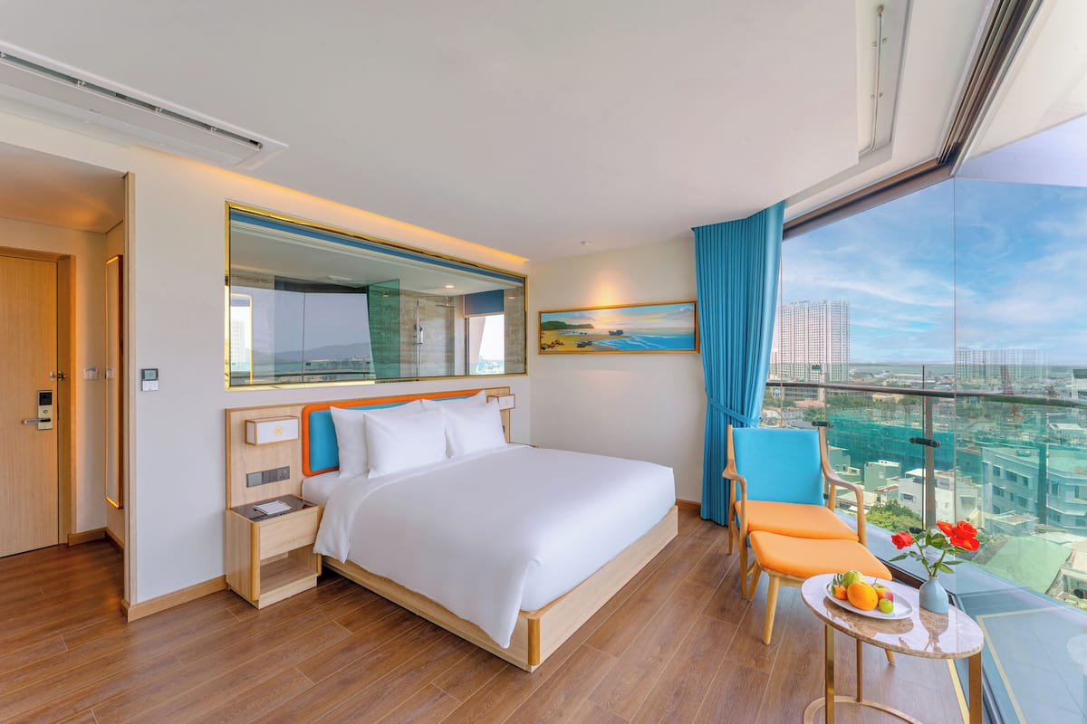 A hotel room with a large bed, a nightstand, a small table with a vase of flowers, a chair, and a view of the city through large windows.