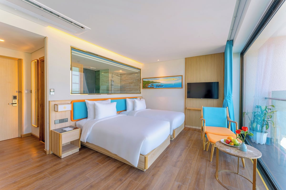 A modern hotel room with a large bed, a bedside table, a small table with fruit, a chair, and a large window with a view.
