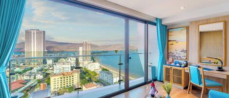 A modern hotel room with a large balcony, a desk, and a view of the city and water.