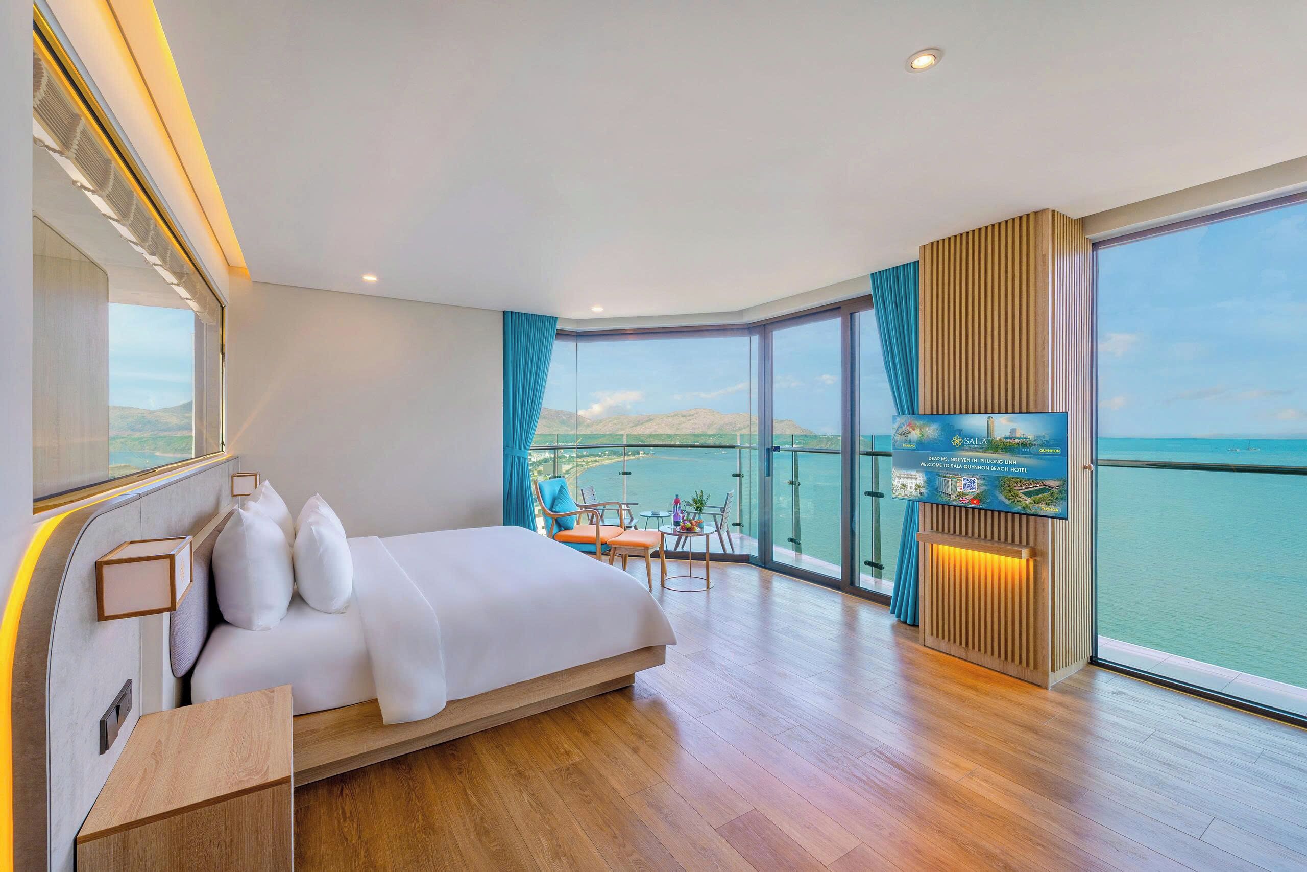 Une chambre d’hôtel moderne avec un grand lit, un balcon offrant une vue sur la mer et une pancarte accueillant les clients à l’hôtel Sala Quynh Hoa Beach.