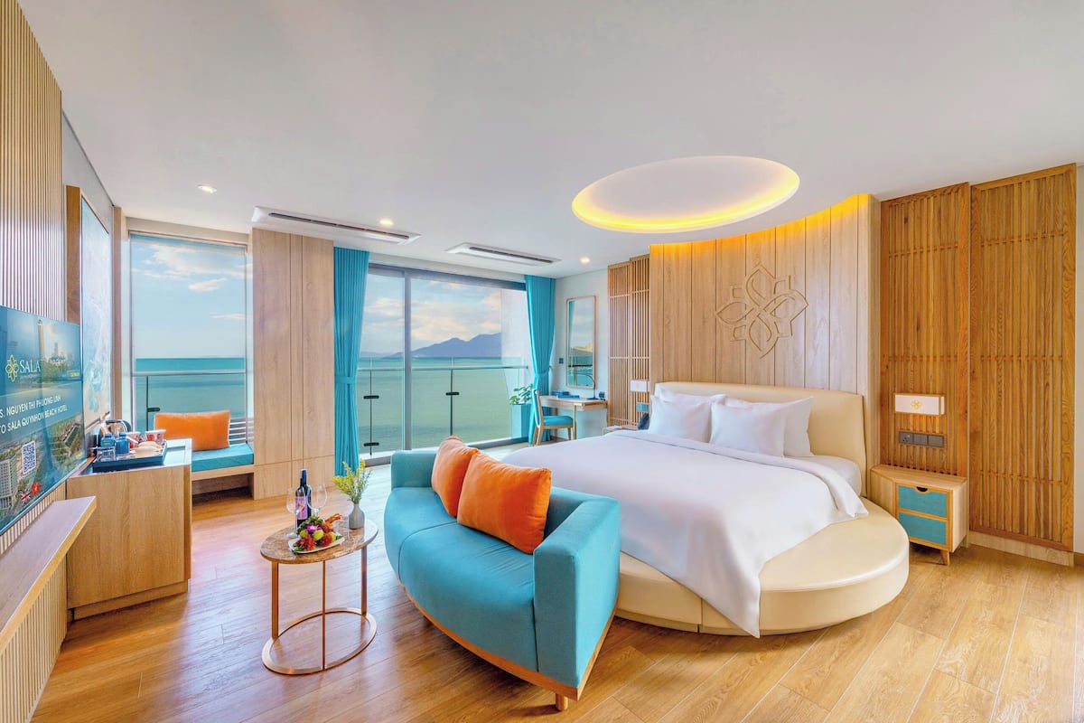 A modern hotel room with a large bed, a sofa, a desk, and a view of the sea.