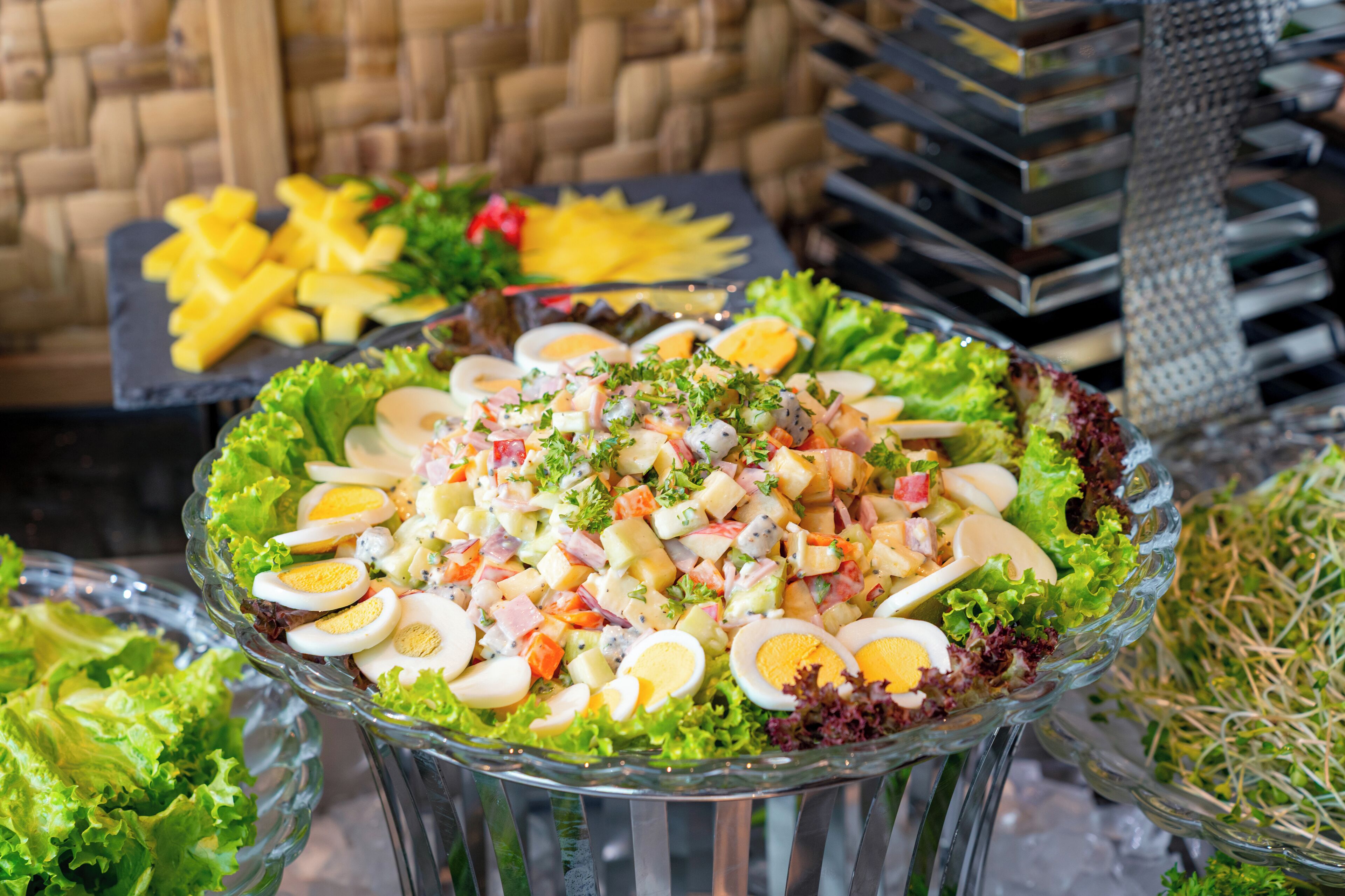 A glass bowl filled with a salad, surrounded by other food items and a wicker basket in the background.