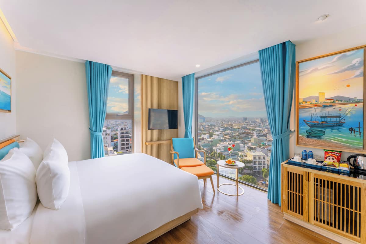 A hotel room with a large bed, a TV, a chair, a small table, and a view of the city.