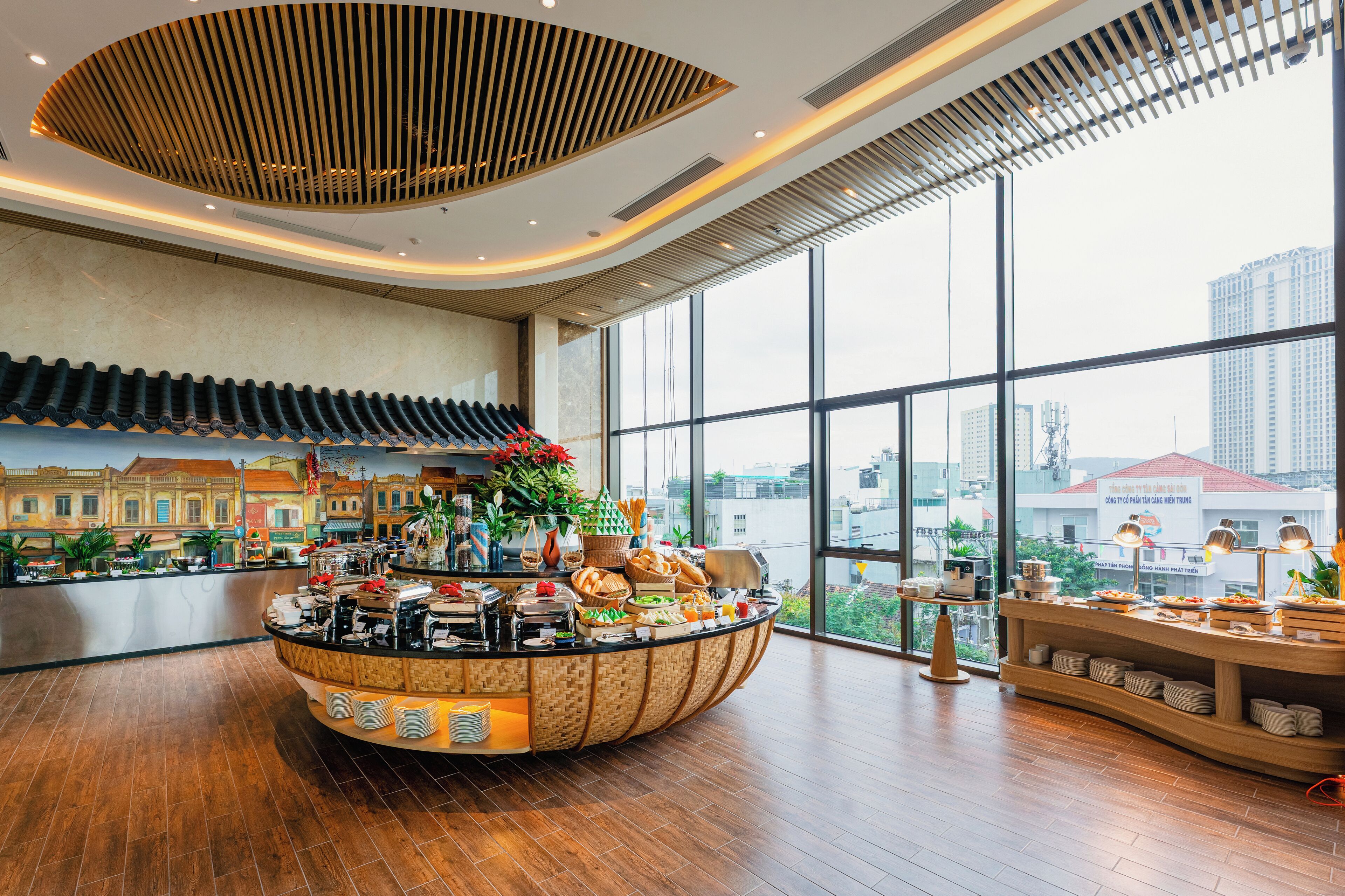 A modern hotel lobby with a central buffet, large windows offering city views, and a traditional architectural element on the ceiling.