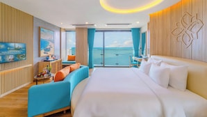 Hotelkamer met een groot bed, een zithoek met een bank en fauteuils, een kleintje tafeltje met een vaas en fruit, een televisie, en uitzicht op de zee door grote ramen.