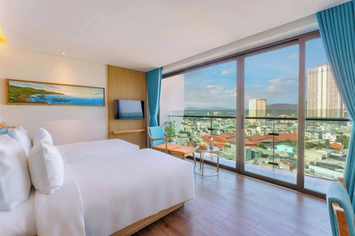 A hotel room with a large bed, a TV, a painting, and a view of the city.