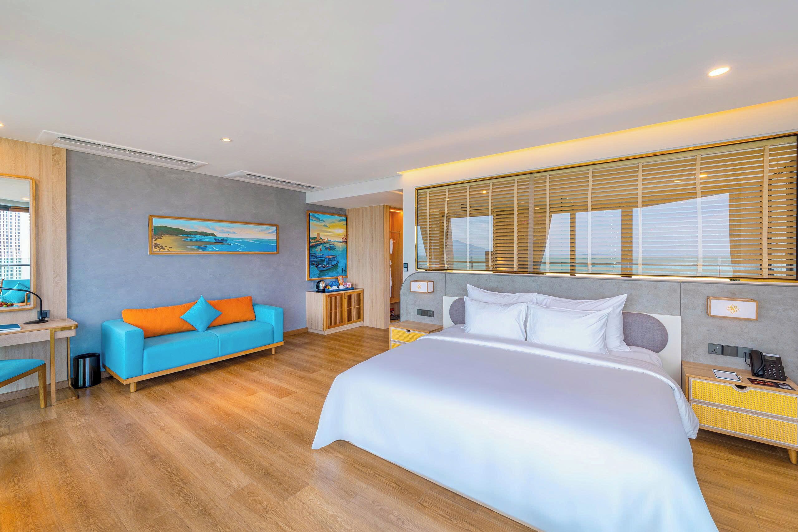 A modern hotel room with a large bed, a sofa, a desk, and a window with blinds.