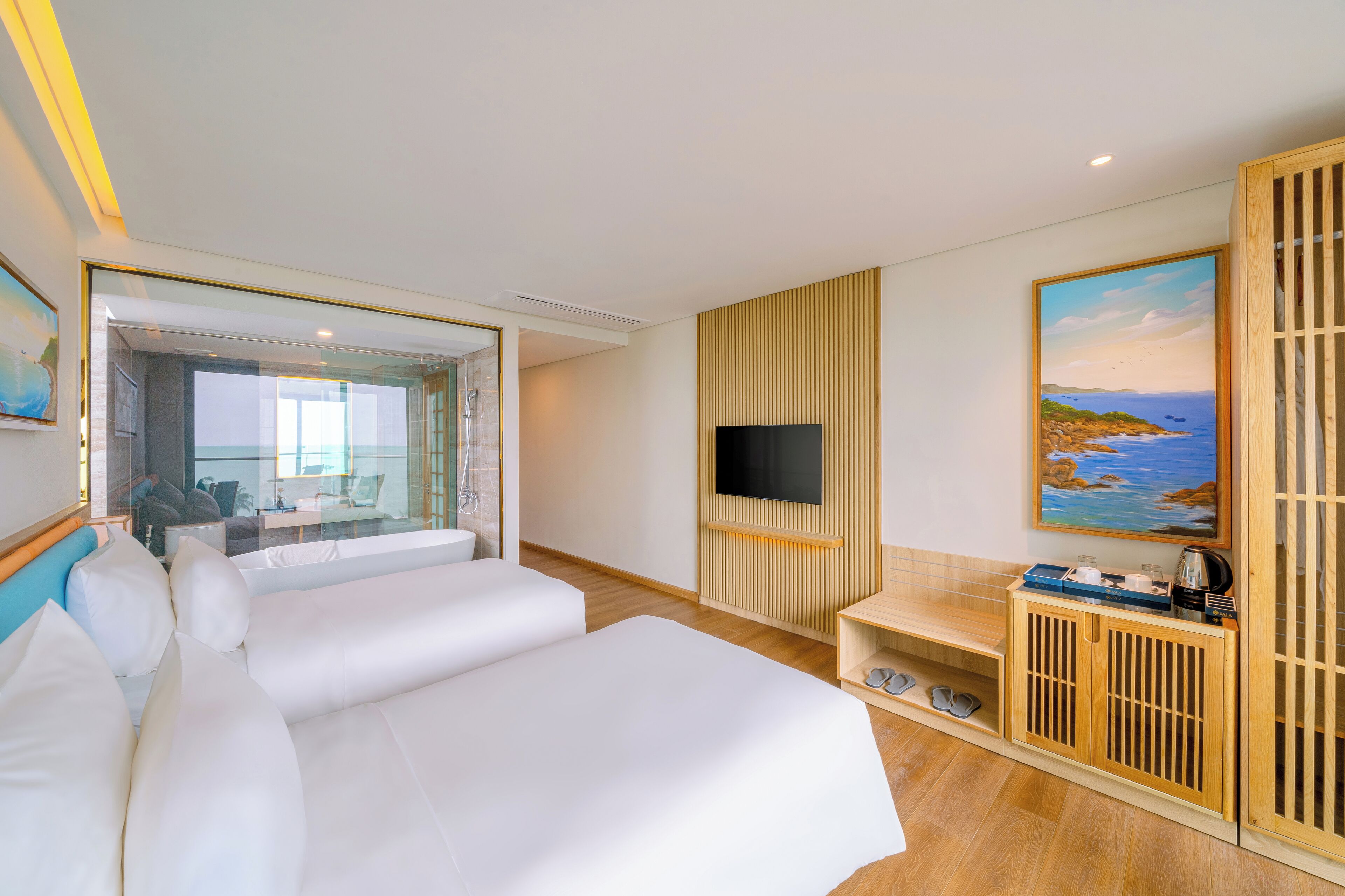 Ett modernt hotellrum med en stor säng, en tv som är monterad på en träpanel, en tavla som föreställer ett kustlandskap och en utsikt över rummets inredning genom en glasdörr.