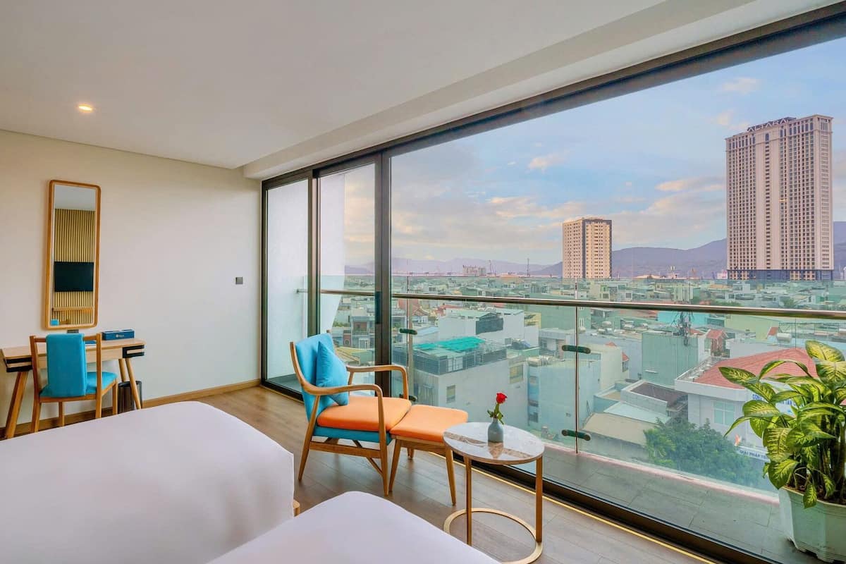 A hotel room with a large window, a desk, two chairs, a bed, and a small table with a vase of flowers.
