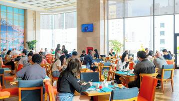 A spacious dining area with multiple tables and chairs, large windows offering a view of the city, and a television mounted on the wall.