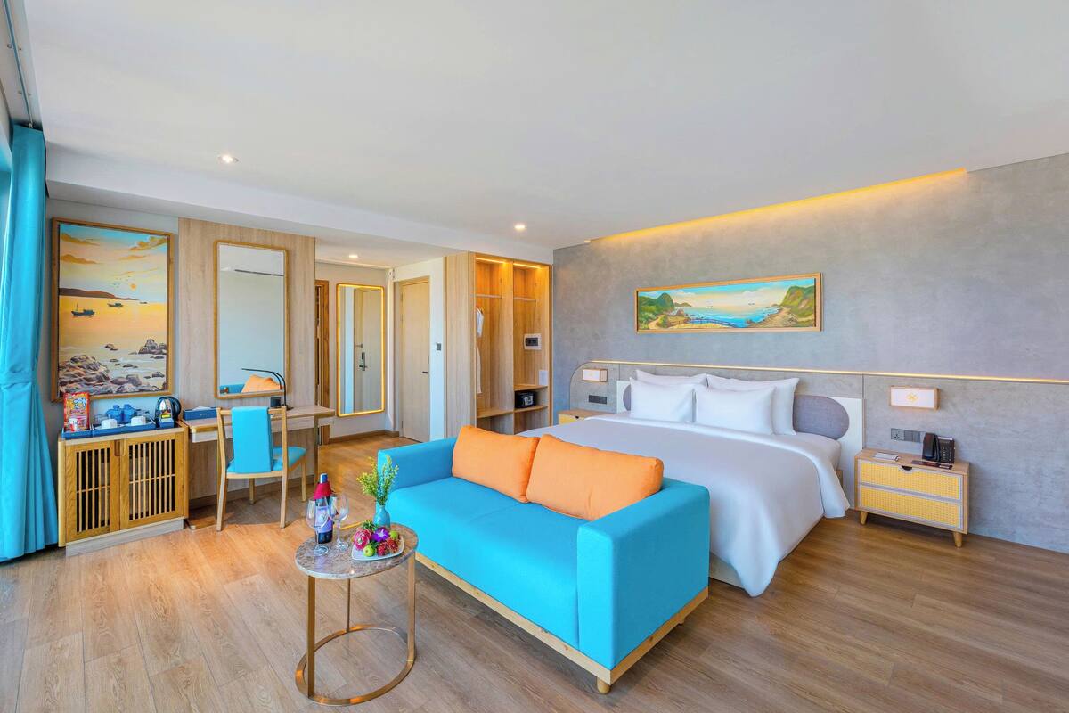 A modern hotel room with a bed, a blue sofa, a small table with drinks, a wardrobe, and a painting on the wall.