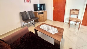 A living room with a sofa, two chairs, a wooden coffee table with a lace doily, a television on a wooden stand, and a door with a red frame. - Apartman Mare 3 (Dramalj)