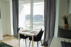 A modern dining area with a white table and black chairs, a vase with a plant, and a view of buildings through large windows. - Bloomfields Studio in Mayan - Yas Island (Abu Dhabi)