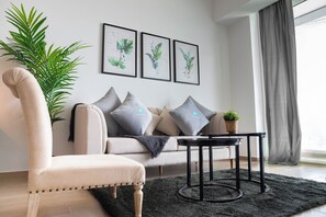 A modern living room with a sofa, armchair, and a coffee table. There are three framed botanical prints on the wall, a potted plant, and a large window with curtains. - Bloomfields Studio in Mayan - Yas Island (Abu Dhabi)
