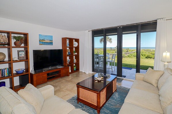 A living room with a TV, wooden shelves, and a view of the ocean through large windows.