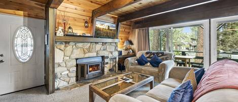Una sala de estar acogedora con chimenea de piedra, techo de madera y grandes ventanales con vistas al exterior.