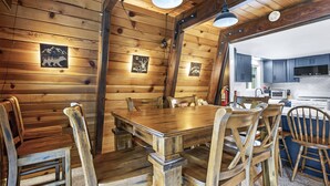 Een houten eettafel met stoelen, een keuken met kastjes, en een plafond met houten balken en verlichting.