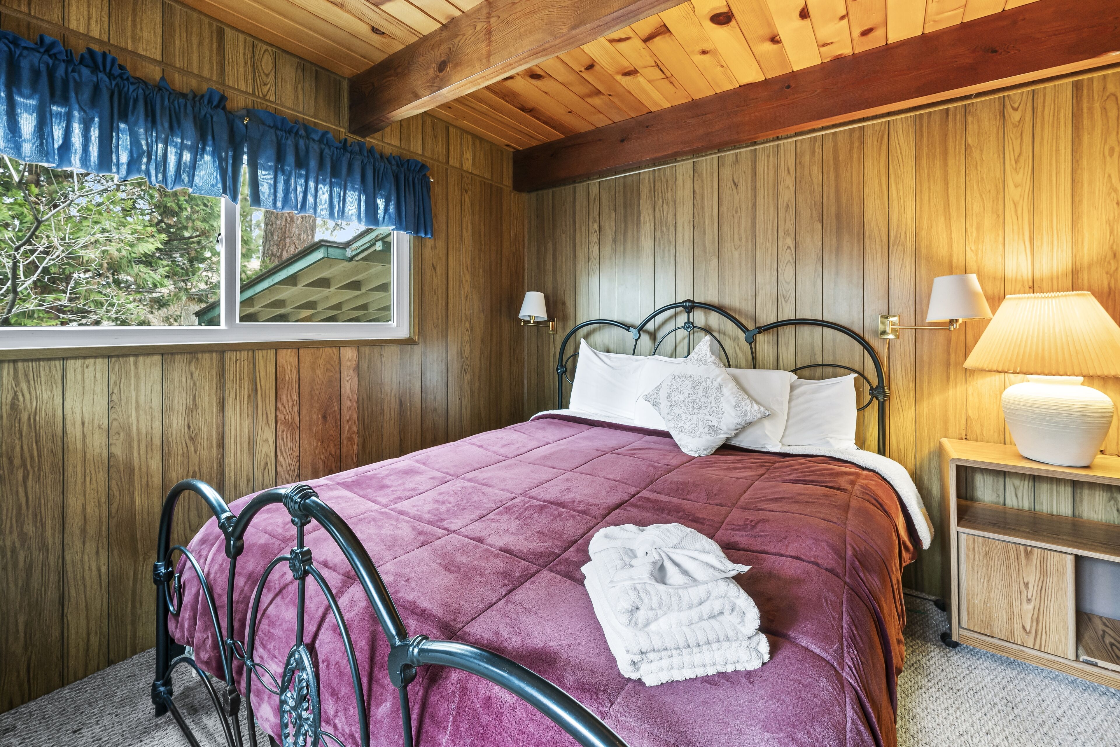 A wooden-paneled bedroom with a bed, two bedside lamps, a window with blue curtains, and a view of trees outside.