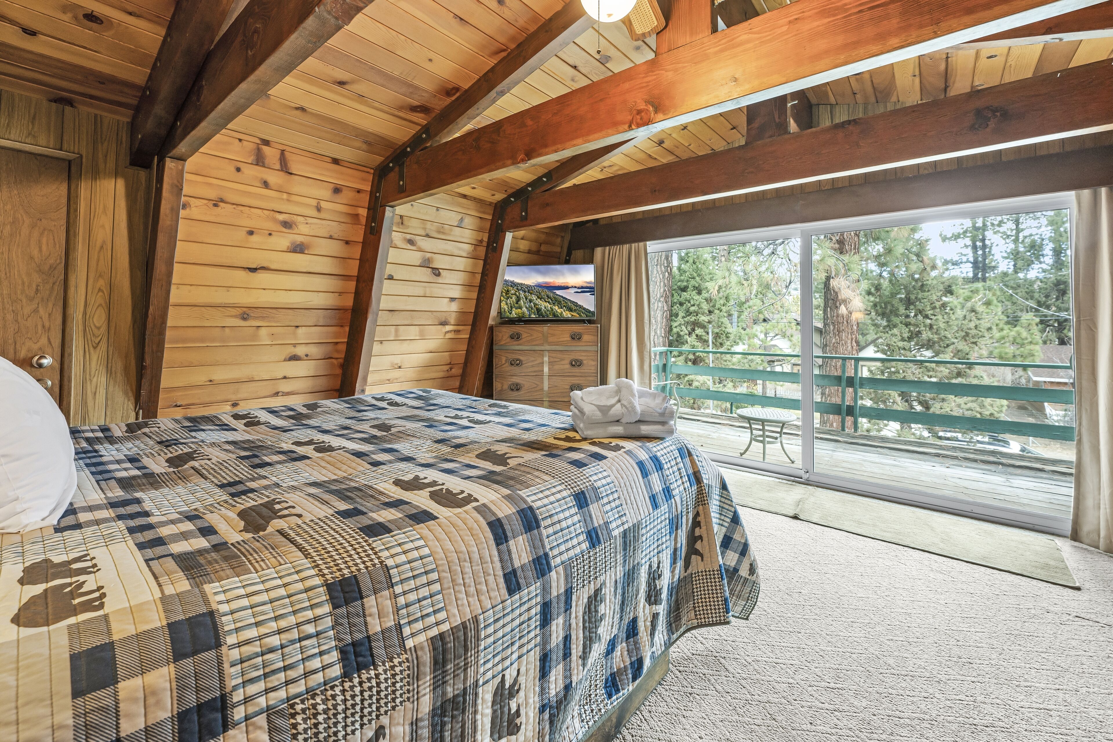 A bedroom with a checkered bedspread, wooden walls, a window with a view of trees, and a balcony with a table and chairs.