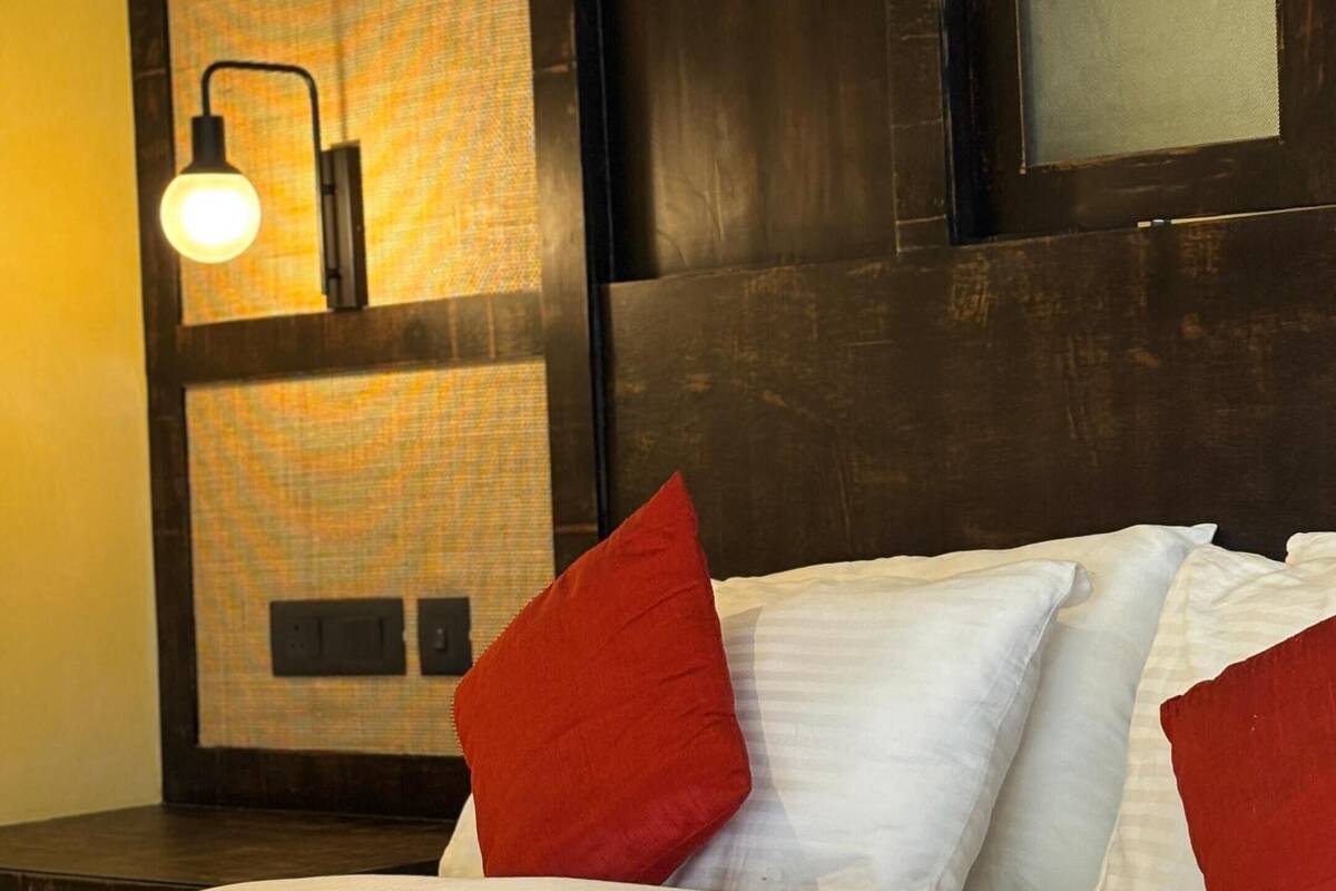 A hotel room with a large bed, white bedding, red pillows, a dark headboard, and a wall-mounted lamp.