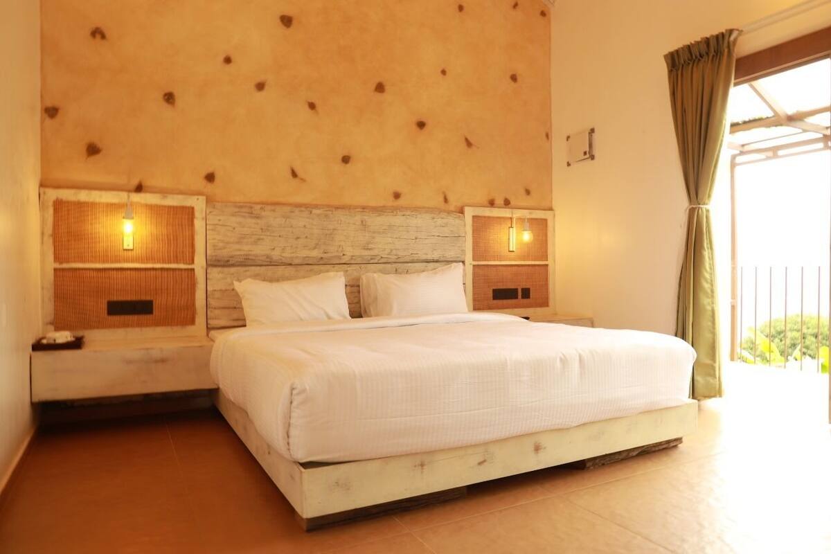 A bedroom with a wooden headboard, two bedside tables with lamps, a bed with white bedding, and a view of greenery outside.