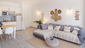 Ein modernes Wohnzimmer mit einem grauen Sofa, einem kleinen Esstisch und einer Küchenzeile im Hintergrund.