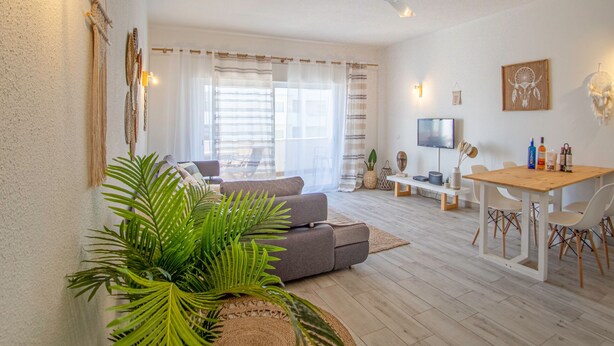 Ein geräumiges Wohnzimmer mit grauem Sofa, einem Holzesstisch mit Stühlen, einem Deckenventilator und einem Fernseher an der Wand.