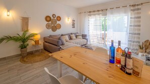 Ein gemütliches Wohnzimmer mit einer Couch, einem Holztisch und einem Essbereich. Es gibt Wanddekorationen und eine Pflanze.