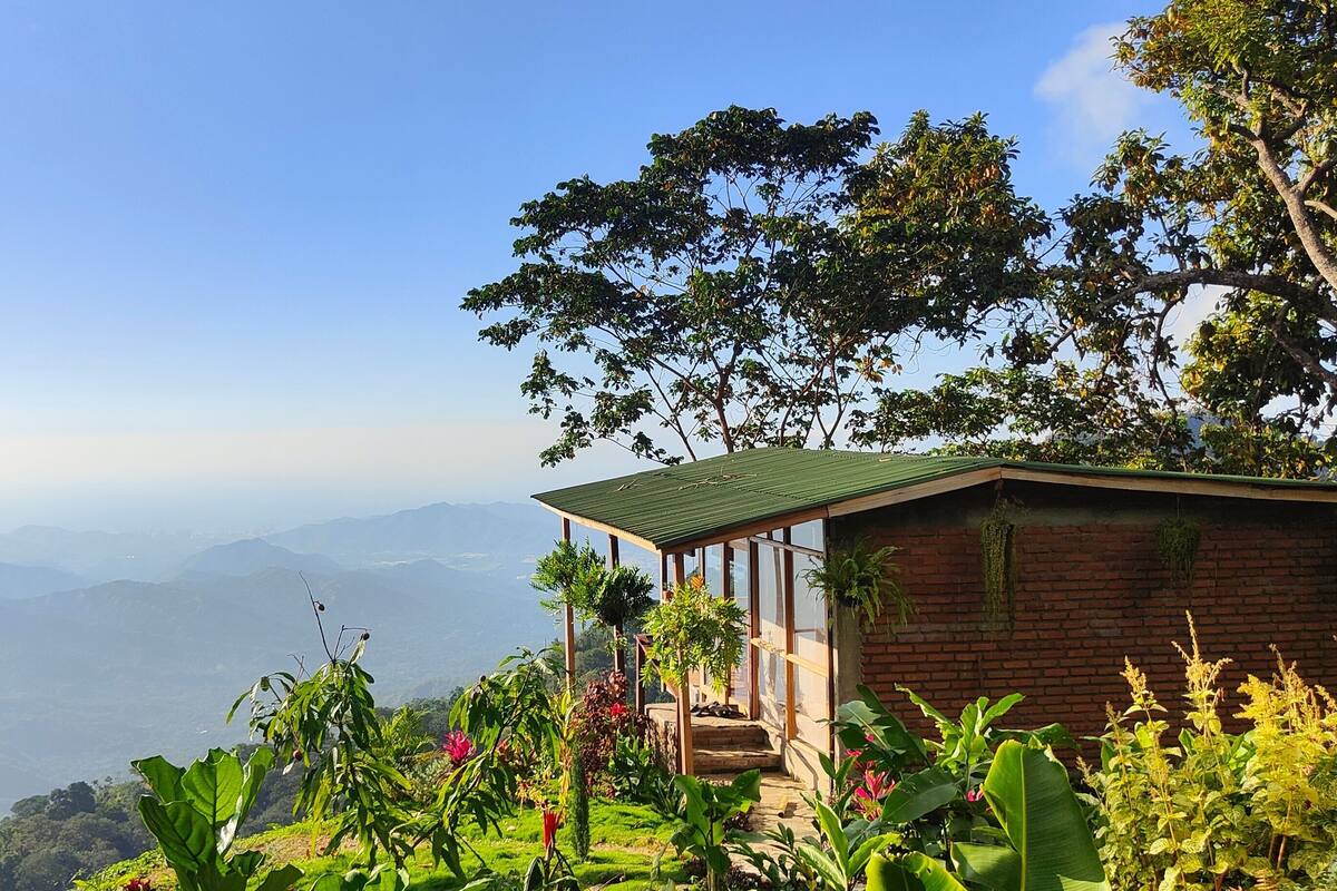 A rustic cabin with a green roof, surrounded by lush greenery and a mountainous backdrop.