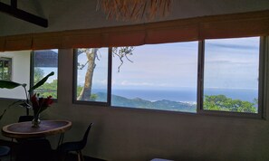 A room with a large window overlooking a coastal landscape, a small round table with chairs, and a potted plant. - Minca Sintropia Organic Coffee Finca (Minca)