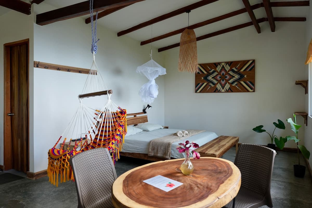 A cozy room with a hammock, a bed, a round wooden table with a vase, and a window with curtains.