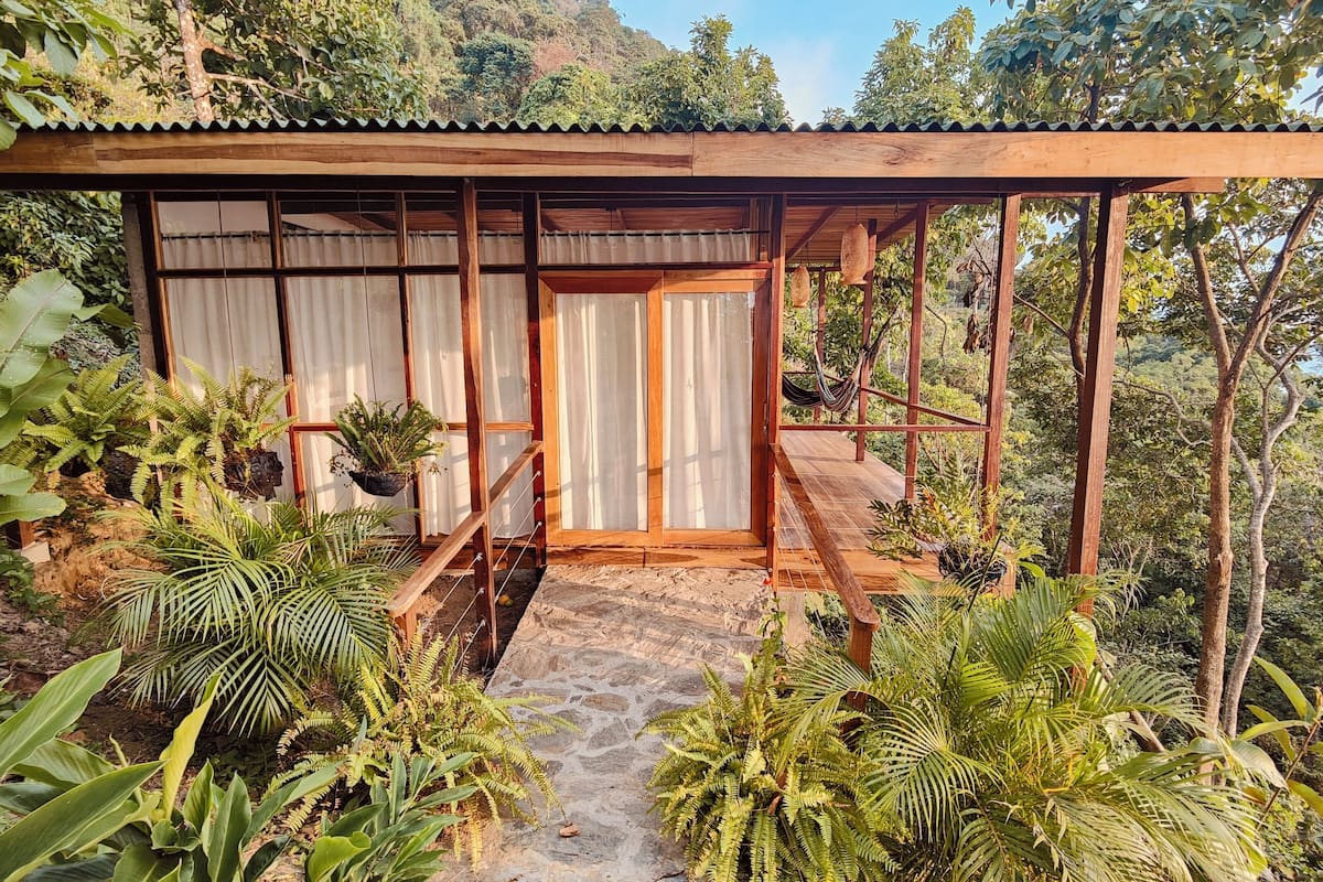 A wooden structure with a covered porch, surrounded by lush greenery and a stone pathway leading to the entrance.