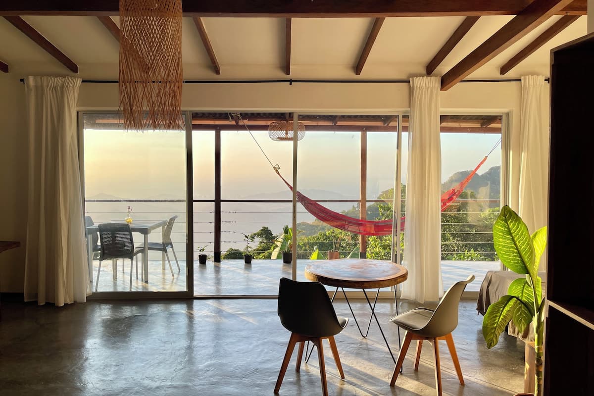 A spacious room with a large sliding glass door leading to a balcony with a view of the sea and mountains. There is a round wooden table with chairs, a hanging chair, and a potted plant.