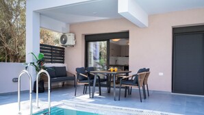A modern outdoor pool area with a dining set and lounge chairs, a built-in air conditioning unit, and a view into the adjacent kitchen.